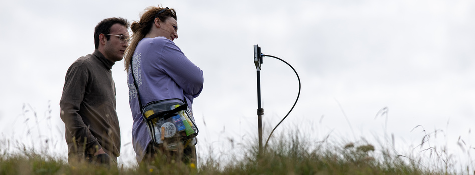 Two people in a field with grass examining equipment mounted on a tall stand.