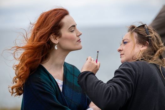 Woman with red hair in profile while another person applies makeup with a brush.