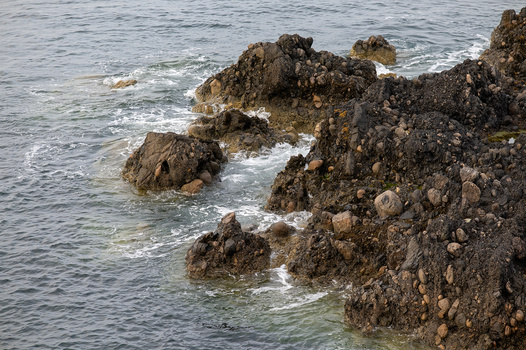 Rocky coastline with weathered stone formations meeting calm ocean water and white foam.