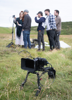 Professional video camera on tripod in foreground with film crew gathered in background on grassy hillside.