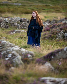 Young woman with long reddish-brown hair standing among rocky outcrops in a moorland landscape.