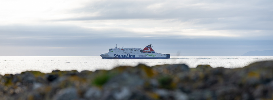 Stena Line ferry ship on calm water with rocky shoreline in sharp foreground blur.