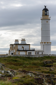 White lighthouse with attached stone keeper's house under dramatic cloudy sky on rocky terrain.