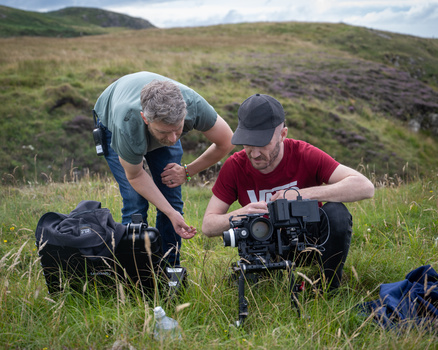 Two photographers in a grassy moorland setting, one crouching while adjusting a professional video camera held by the other in a blue shirt.