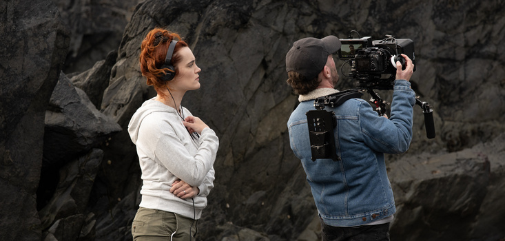 Cinematographer operating a professional cinema camera on a tripod while filming a subject against a textured stone wall.