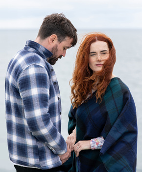 Man in blue plaid shirt looking down beside woman with red hair in dark sweater against ocean backdrop.