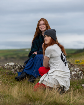 Two young women posing together on a grassy hillside with a soft, overcast sky in the background.