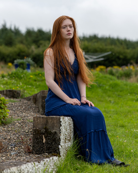 Woman with long reddish hair sitting on a stone block in a garden setting, wearing a dark blue dress.