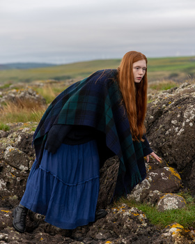 Red-haired woman in navy dress leaning against moss-covered stone in rolling countryside landscape.