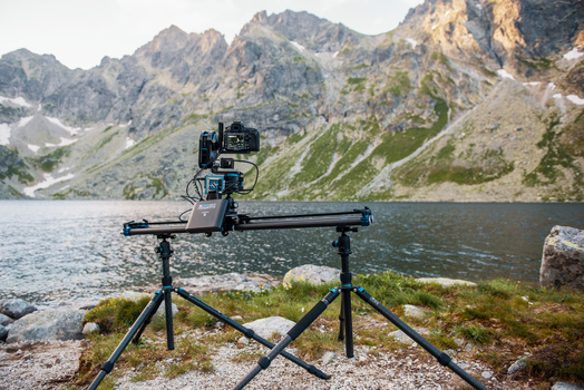 Camera slider rig mounted on tripod legs positioned at the shoreline of a mountain lake.