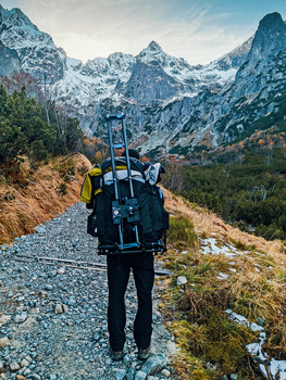 Backpacker with large hiking pack walking on gravel path toward snow-capped mountain peaks.