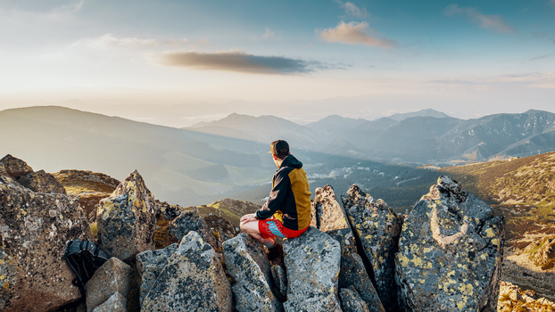 Hiker in yellow jacket sitting on mountain rocks overlooking layered valley and distant peaks.