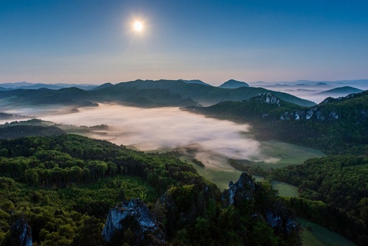 Forested mountains rise above a thick blanket of morning fog under a bright sun in a clear blue sky.