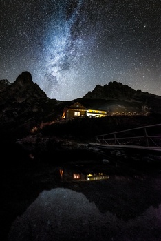 Milky Way arcs across night sky above illuminated building nestled between dark mountain silhouettes.