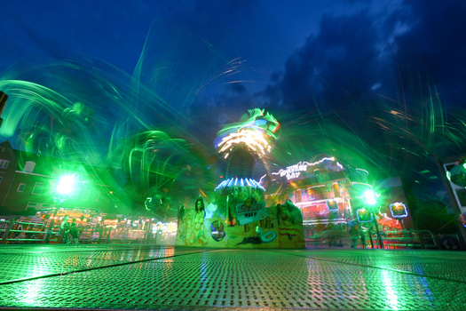 Nighttime fairground with green and blue neon lighting illuminating carnival rides and structures.