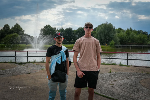 Two people standing on sandy ground with a fountain and trees visible across a body of water behind them.