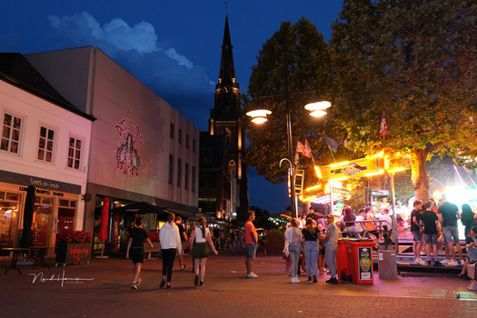 Evening street scene with crowds, illuminated church spire, and warm-toned vendor stalls under blue twilight sky.