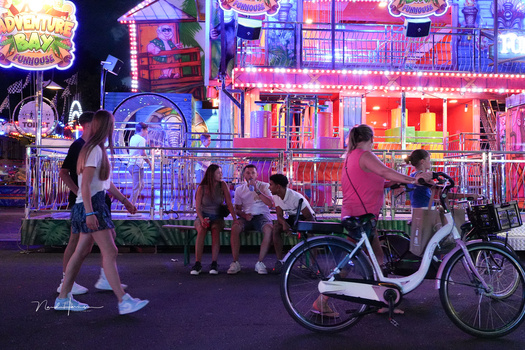 Nighttime carnival scene with neon-lit game booths, people walking and riding bikes on dark pavement.