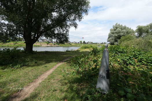 Numbered marker post along a dirt path beside a river, with trees and green landscape in background.