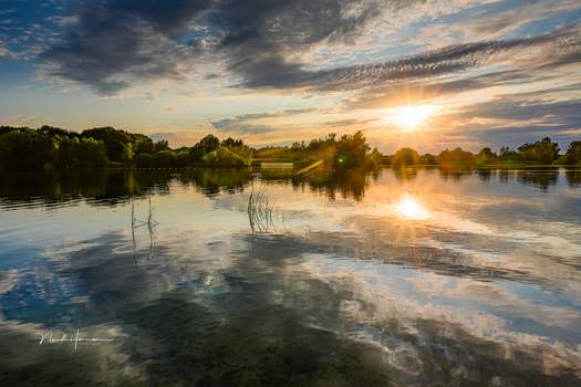 Calm river at sunset with trees reflected in still water under dramatic clouds.
