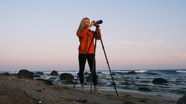 Photographer with long hair in orange jacket using tripod-mounted telephoto lens on beach at dusk.