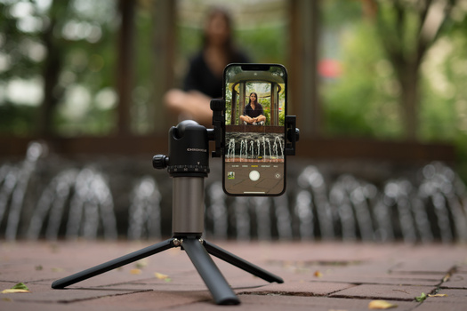 Smartphone mounted on tripod displaying portrait of woman, with blurred figure in background outdoors.