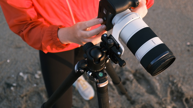 Photographer in red jacket holding mirrorless camera with white telephoto lens on tripod.