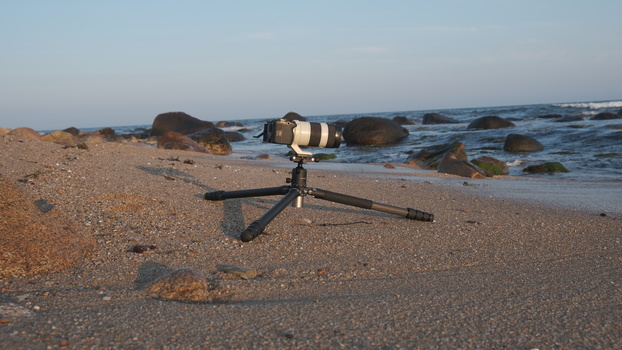 Telephoto lens mounted on tripod on sandy beach with rocky coastline and ocean in background.
