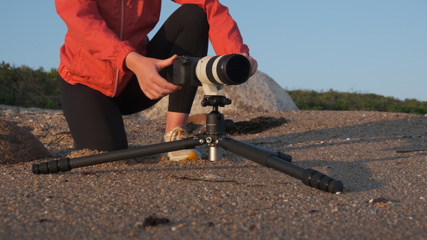 Photographer in red jacket adjusting a telephoto lens mounted on a tripod at ground level.