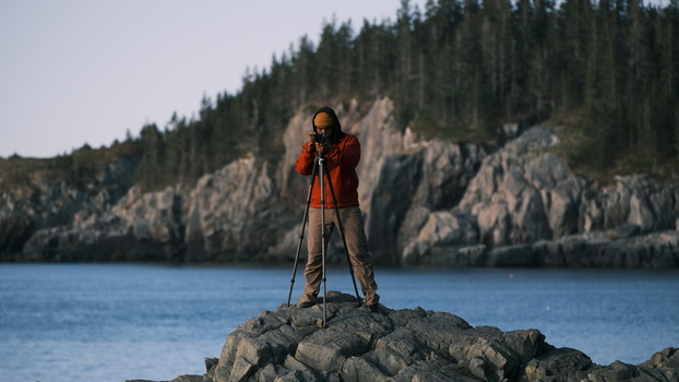 Hiker in red jacket stands on rocky outcrop overlooking calm blue lake with forested cliffs.