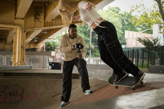 Skateboarder performing an aerial trick under a concrete bridge while another person films with a camera.