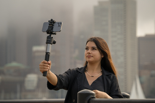 Woman holding a selfie stick with smartphone against a blurred cityscape backdrop.