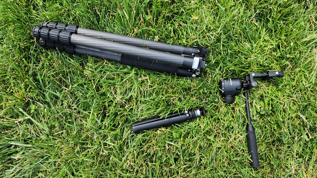 Three pieces of camera support equipment laid out on grass: a tripod, ball head, and center column.