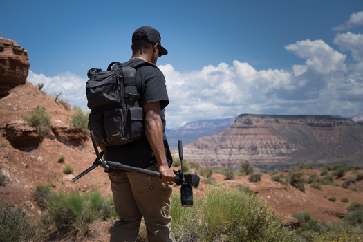 Hiker with large backpack and trekking poles stands on rocky terrain overlooking a vast canyon landscape.