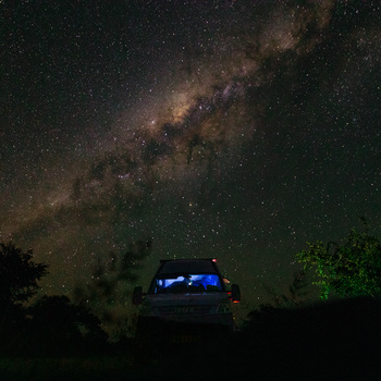 Milky Way galaxy stretching across night sky above a vehicle with interior lights on, surrounded by silhouetted trees.
