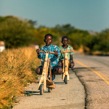 Two young people riding bikes on a paved rural road with dry grassland on both sides.