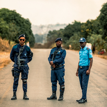 Three people standing in the center of a straight road lined with trees, wearing blue uniforms and caps.