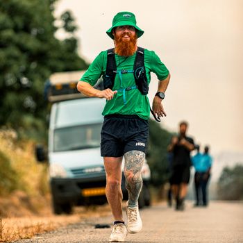 Male runner in green athletic gear and hat jogging on a dusty road with spectators and vehicles in the background.