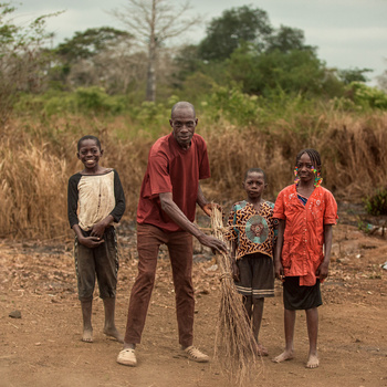 An adult and three children stand together on a dirt path holding bundled dried grass and branches.