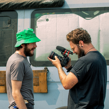 Cinematographer filming a bearded man wearing a green cap against a light blue wall.