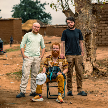 Three men posing outdoors in an arid setting with adobe structures and trees visible in the background.