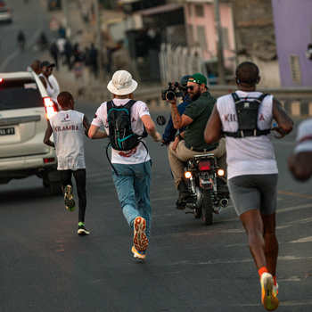 Runners in white shirts sprint down an urban street alongside a motorcyclist.
