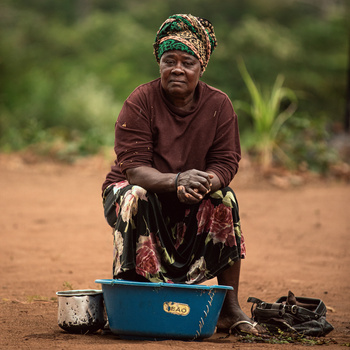 Woman in maroon clothing and patterned headwrap sits on ground beside blue basin and cooking vessels.