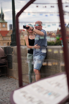 Man helping woman adjust camera settings on rooftop with historic cityscape and clock tower in background.