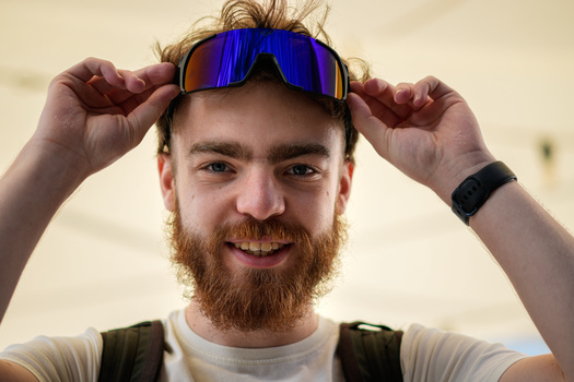 Man with reddish beard holding blue-mirrored sports sunglasses on forehead, smiling at camera.