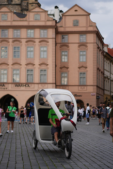 Cyclist riding a white cargo bike through a European town square with historic terracotta buildings in the background.