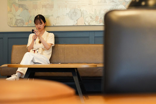 Woman in cream-colored clothing sitting on a tan leather bench against a blue-green paneled wall.