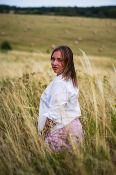 Woman in white shirt standing in tall grass field, looking over shoulder at camera.