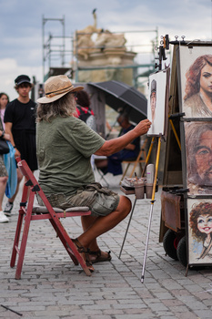 Street artist painting a portrait on canvas while seated on a red stool in a public plaza.