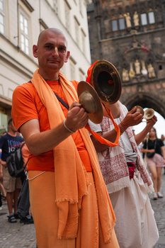 Monk in orange robes playing traditional hand cymbals on a city street with historic architecture in background.